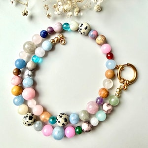 Puede incluir: Un collar de cuentas de colores con una variedad de cuentas de piedras preciosas en tonos rosa, azul, verde y marrón. El collar tiene un cierre dorado y se muestra sobre una superficie blanca.