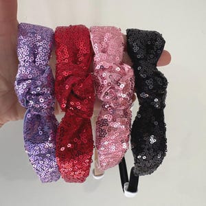 May include: Four sequined headbands in purple, red, pink, and black. Each headband features a bow design and is covered in small, shiny sequins. The headbands are held together, showcasing the variety of colors and styles.