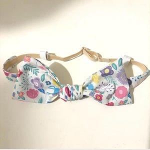 May include: A floral bow tie with an adjustable beige strap. The bow tie features a white background with a pattern of pink, purple, yellow, and blue flowers and green leaves. The bow tie is designed for a comfortable fit.