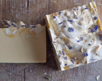 May include: Two bars of handmade soap. One is a solid light yellow color. The other is white with blue and yellow floral accents. The soap is cut into bars and sits on a wooden surface.