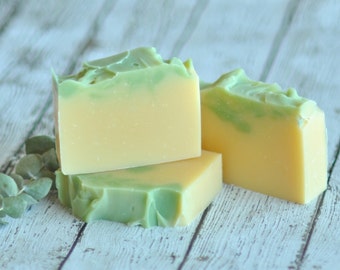 May include: Three bars of handmade soap with a green and yellow swirl design. The soap is on a white wooden surface.