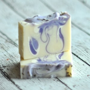 May include: Two bars of handmade soap with a swirl design in shades of white and purple. The soap is made with lavender and has a natural, earthy scent.
