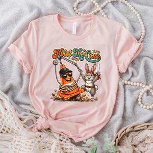 Puede incluir: Camiseta rosa claro con un gr&aacute;fico de dibujos animados y el texto "Hoist The Cone". El gr&aacute;fico representa un cono de tr&aacute;fico naranja con una gorra de b&eacute;isbol y un conejo de dibujos animados con un cuchillo. El dise&ntilde;o es de estilo retro.