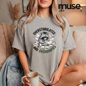 Könnte beinhalten: Graues T-Shirt mit der Aufschrift "OVERSTIMULATED AND OVERQUALIFIED" und einem Cartoon-Waschbären mit Doktorhut, umgeben von Kaffeetassen und Papieren.