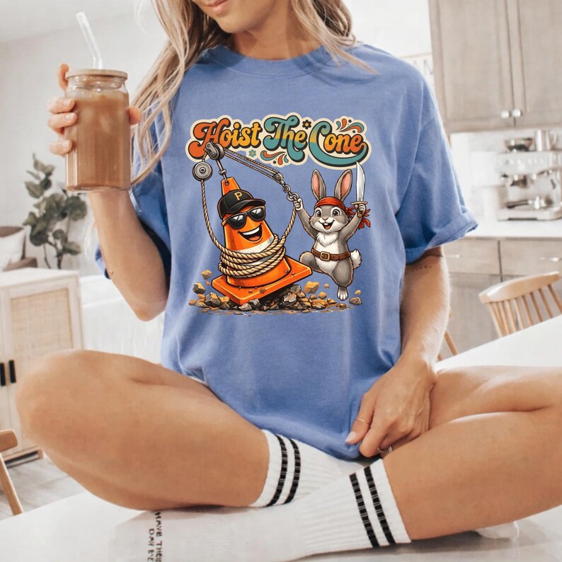Puede incluir: Camiseta azul claro con el texto "Hoist The Cone" en una fuente retro. El gr&aacute;fico presenta un cono de tr&aacute;fico de dibujos animados con gafas de sol y una gorra de b&eacute;isbol, izado por un conejo pirata. El cono est&aacute; envuelto en una cuerda.