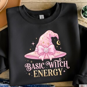 Pode incluir: Camisola preta com um chapéu de bruxa rosa com laço, estrelas e lua crescente. O texto "BASIC WITCH ENERGY" está impresso abaixo. O design está centrado na frente da peça de vestuário.