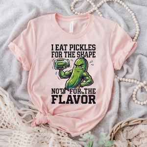 May include: Pale pink t-shirt with a cartoon pickle design. The green pickle character is flexing its muscles and holding a jar, with the text "I EAT PICKLES FOR THE SHAPE NOT FOR THE FLAVOR". The shirt is made of a soft material.