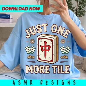 May include: A light blue t-shirt featuring a large graphic of a mahjong tile with the red Chinese character for middle, surrounded by smaller tiles and coins. The text "JUST ONE MORE TILE" is printed in a bold, arched font above and below the tile.
