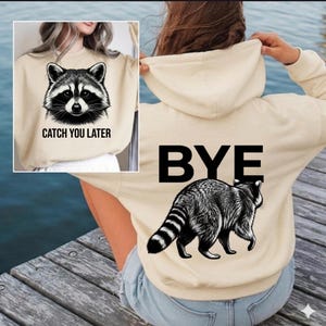 Puede incluir: Sudadera con capucha beige con un diseño de mapache en blanco y negro. La parte delantera dice "CATCH YOU LATER", mientras que la parte trasera tiene "BYE" encima de la ilustración del mapache. Prenda casual.