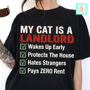 May include: A black t-shirt featuring white and red text that reads MY CAT IS A LANDLORD. Below the text is a checklist with green checkmarks next to the phrases Wakes Up Early, Protects The House, Hates Strangers, and Pays ZERO Rent.