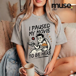 Puede incluir: Una camiseta gris con el texto "I PAUSED MY MOVIE TO BE HERE" y un dibujo de una persona con un mando a distancia y palomitas de maíz. La camiseta es de estilo casual.
