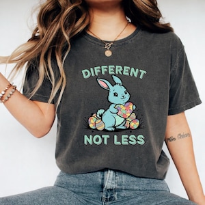 Könnte beinhalten: Dunkelgraues T-Shirt mit einem hellblauen Hasen, der ein herzförmiges Puzzle hält. Der Text "DIFFERENT NOT LESS" befindet sich über und unter der Hasengrafik. Das T-Shirt hat einen Vintage-Look.