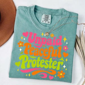 Puede incluir: Una camiseta Comfort Colors de color verde azulado con las palabras "Unpaid Peaceful Protester" en una fuente retro. El dise&ntilde;o incluye texto rosa, naranja y verde con gr&aacute;ficos de flores y corazones. Un collar plateado con un colgante est&aacute; sobre la camiseta.