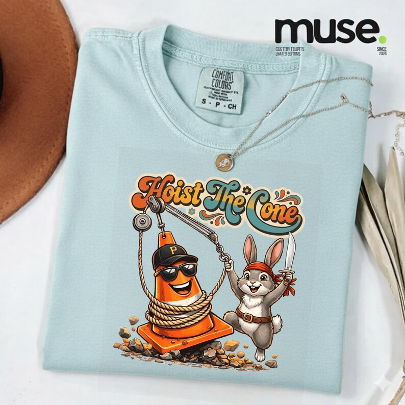 Puede incluir: Camiseta azul claro con el texto "Hoist The Cone" y un gr&aacute;fico de un cono de tr&aacute;fico naranja sonriente con gafas de sol y gorra negra, y un conejo con una espada. La camiseta es de la marca Comfort Colors.