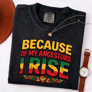 Può includere: T-shirt grigio scuro con la scritta "BECAUSE OF MY ANCESTORS I RISE" in giallo, rosso e verde. Una collana con ciondolo a stella e un orologio in pelle marrone sono visibili.