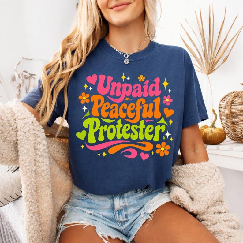 Puede incluir: Camiseta azul marino con la frase "Unpaid Peaceful Protester" en un estilo retro. El dise&ntilde;o incluye flores, corazones y estrellas en rosa, naranja y verde. La camiseta es de estilo casual.