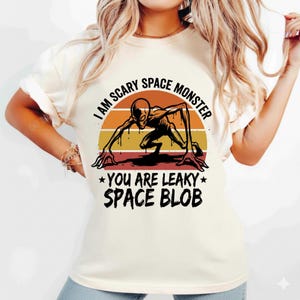 May include: Cream-colored t-shirt featuring a graphic of a skeletal alien creature in black, orange, and yellow. The text reads "I AM SCARY SPACE MONSTER" and "YOU ARE LEAKY SPACE BLOB".