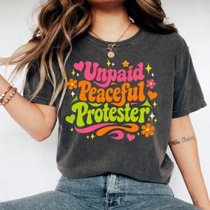 Puede incluir: Camiseta gris oscuro con la frase "Unpaid Peaceful Protester" en una fuente retro. El dise&ntilde;o incluye texto rosa, naranja y verde con motivos de flores y corazones. Una prenda de vestir informal.