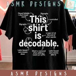 Op de afbeelding: Een zwart t-shirt met witte tekst die This shirt is decodable leest. Het ontwerp bevat diverse taalkundige annotaties met pijlen die naar specifieke woorden wijzen, waarbij concepten zoals gesloten lettergrepen en tweeklanken worden uitgelegd.