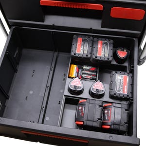Può includere: Un cassetto portautensili in plastica nera con maniglia rossa, dotato di uno scomparto aperto a sinistra e una sezione suddivisa a destra contenente varie batterie rettangolari e circolari per utensili elettrici e piccole pile cilindriche.