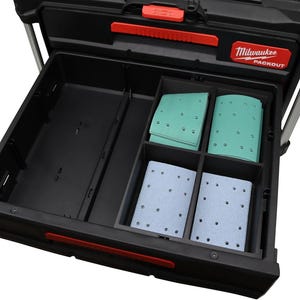 Puede incluir: Caja de herramientas negra Milwaukee Packout con asa y logotipo rojos. El cajón abierto contiene varios compartimentos con almohadillas de lijado verdes y azul claro. La caja de herramientas está diseñada para la organización y el almacenamiento.
