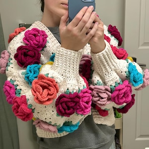 Op de afbeelding: Een wit gebreid vest met kleurrijke bloemenversieringen. Het vest heeft een V-hals en lange mouwen.