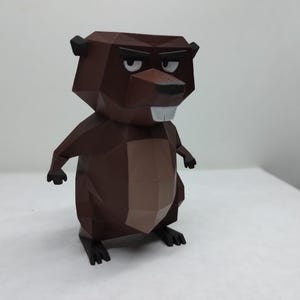 Puede incluir: Una figura de castor de papel marrón con una expresión severa. El diseño geométrico presenta una cabeza y un cuerpo de color marrón oscuro, con acentos marrón claro en el pecho y el vientre. El castor tiene pies negros y orejas pequeñas.
