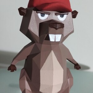 Puede incluir: Una figura geométrica de castor de estilo low-poly con una gorra de béisbol roja. El castor tiene una expresión severa, con dientes blancos y una nariz marrón. La figura está pintada en tonos marrones y beige.