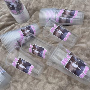 Peut inclure: Gobelets en plastique transparents avec un autocollant rose indiquant "Wife of the party". Les gobelets sont &eacute;parpill&eacute;s sur une surface textur&eacute;e brun clair. Certains gobelets sont empil&eacute;s.