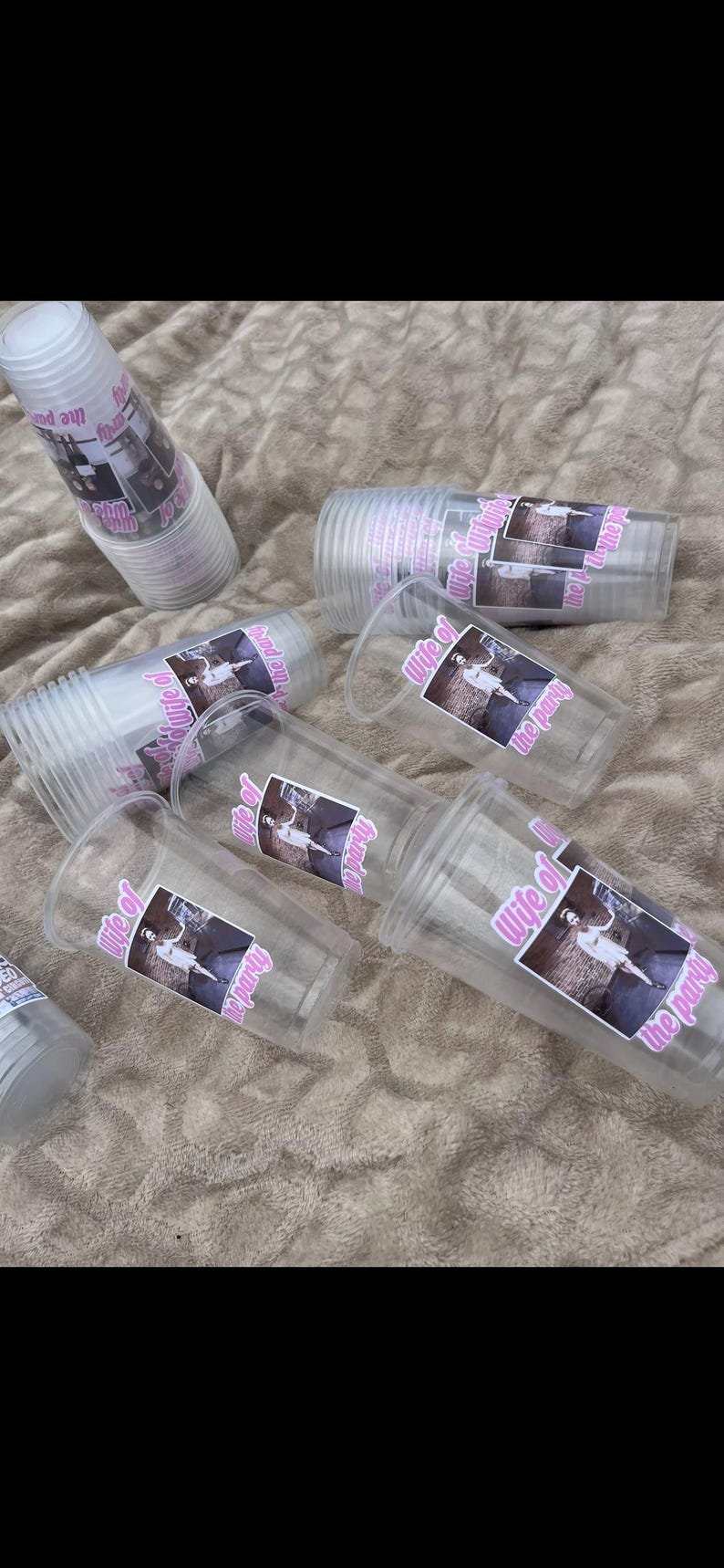 Peut inclure: Gobelets en plastique transparents avec un autocollant rose indiquant "Wife of the party". Les gobelets sont &eacute;parpill&eacute;s sur une surface textur&eacute;e brun clair. Certains gobelets sont empil&eacute;s.