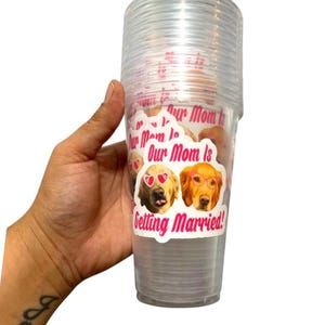 Peut inclure: Une pile de gobelets en plastique transparents avec un autocollant indiquant "Our Mom Is Getting Married!". L'autocollant repr&eacute;sente deux chiens golden retriever portant des lunettes de soleil et des lunettes. Les gobelets sont tenus dans une main.