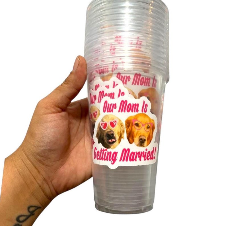 Peut inclure: Une pile de gobelets en plastique transparents avec un autocollant indiquant "Our Mom Is Getting Married!". L'autocollant repr&eacute;sente deux chiens golden retriever portant des lunettes de soleil et des lunettes. Les gobelets sont tenus dans une main.