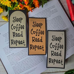 以下が含まれることがあります： 「Sleep Coffee Read Repeat」の文字が黒い筆記体で書かれた、長方形のブックマークが3つ。ブックマークはベージュ、タン、濃いグレーの色合いで、ハートのシンボルが「love」の代わりに使われています。開いた本の上に置かれています。