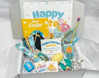 May include: A white box filled with Easter-themed gifts and decorations. The box contains a variety of items, including a card that says "Happy Easter Austin", a small chalkboard, a bunny-shaped pencil, a small notebook, a plastic egg, and a few other items.