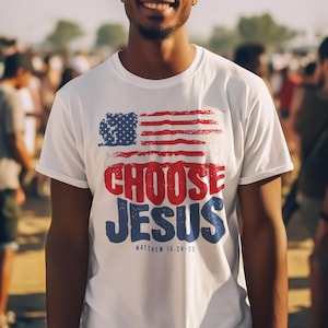 Könnte beinhalten: Weißes T-Shirt mit einem abgenutzten amerikanischen Flaggen-Design und dem Text "Choose Jesus" in roten und blauen Buchstaben. Der Text "Matthew 16:24-25" ist unter dem Design gedruckt.