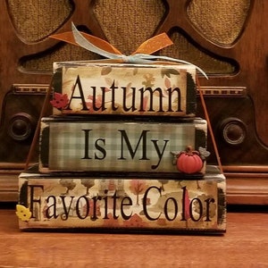 May include: A three-tiered wooden block sign with the text "Autumn Is My Favorite Color". Each block has a different autumn-themed pattern and is decorated with small fabric embellishments.