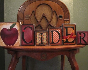 May include: Wooden blocks spelling out the word "CIDER" with a red apple illustration on the first block. The blocks are decorated with fall-themed patterns like polka dots, leaves, and plaid.