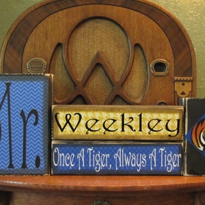 May include: Three wooden blocks stacked on top of each other. The top block is black with a blue and orange tiger graphic. The middle block is yellow with black and white checkered pattern and the text "Weekley". The bottom block is blue with white text that reads "Once A Tiger, Always A Tiger". The bottom block is next to a blue and black block with the text "Mr."