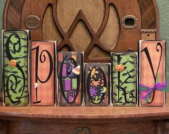 May include: Spooky wooden block letters with a Halloween theme. The letters are decorated with bats, pumpkins, and other Halloween imagery. The letters are arranged in a row on a wooden table.