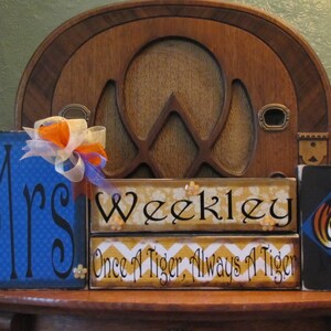 May include: Three wooden signs with a blue, yellow, and black background. The first sign says "Mrs." in black cursive lettering on a blue background with a white and blue checkered pattern. The second sign says "Weekley" in black cursive lettering on a yellow background with a yellow and white chevron pattern. The third sign says "Once a Tiger, Always a Tiger" in black lettering on a yellow background with a yellow and white chevron pattern. The third sign also has a black silhouette of a tiger's head on a blue background.