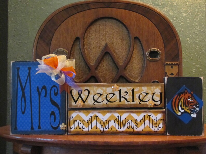 May include: Three wooden signs with a blue, yellow, and black background. The first sign says "Mrs." in black cursive lettering on a blue background with a white and blue checkered pattern. The second sign says "Weekley" in black cursive lettering on a yellow background with a yellow and white chevron pattern. The third sign says "Once a Tiger, Always a Tiger" in black lettering on a yellow background with a yellow and white chevron pattern. The third sign also has a black silhouette of a tiger's head on a blue background.