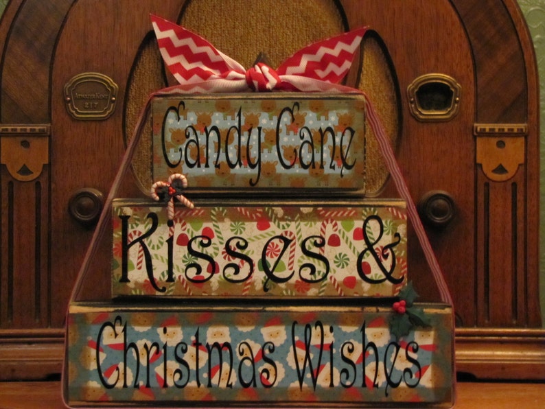 May include: A three-tiered wooden Christmas decoration with a red and white chevron ribbon. The top tier reads "Candy Cane", the middle tier reads "Kisses &", and the bottom tier reads "Christmas Wishes".