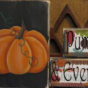 Puede incluir: Un letrero de madera negro con una calabaza naranja pintada y enredaderas verdes. El letrero dice "Pumpkin & Everything Nice".