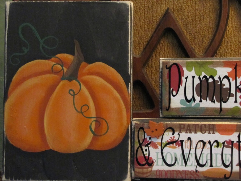 Puede incluir: Un letrero de madera negro con una calabaza naranja pintada y enredaderas verdes. El letrero dice "Pumpkin & Everything Nice".