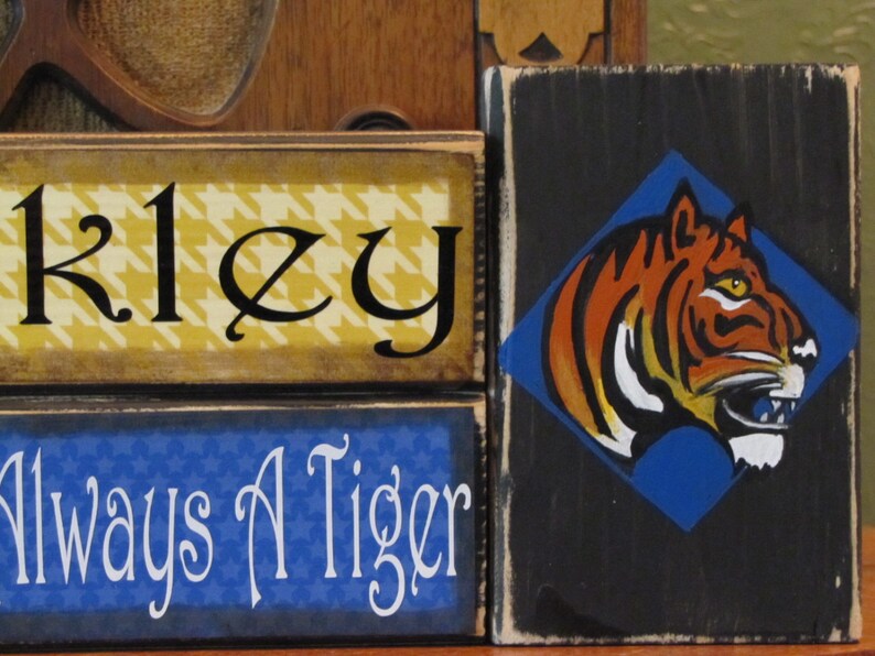 May include: Three wooden signs stacked on top of each other. The top sign is yellow with a black and white houndstooth pattern and the name "Kiley" written in black cursive. The middle sign is blue with the words "Always A Tiger" written in white cursive. The bottom sign is black with a blue diamond shape containing a tiger head illustration. The tiger is orange and black with white stripes and a white mouth.
