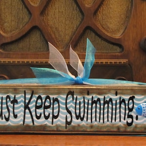 Pode incluir: Uma placa retangular de madeira com a frase "Just Keep Swimming" em escrita preta. A placa tem um padrão de ondas azuis e brancas e um pequeno peixe azul. Uma fita azul e branca está amarrada no topo.