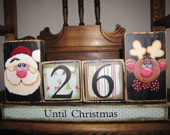 Könnte beinhalten: Ein hölzerner Countdown-Block-Satz für Weihnachten mit einem Weihnachtsmann, einem Rentier und den Zahlen 26. Die Blöcke sind in Schwarz, Weiß, Rot und Grün bemalt. Die Blöcke stehen auf einem Holzregal mit einem Schild, auf dem "Bis Weihnachten" steht.