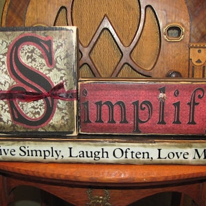 May include: Three wooden blocks stacked on top of each other. The top block is red with black lettering that says "Simplify". The middle block is brown with a gold and black pattern and a black letter "S". The bottom block is brown with white lettering that says "Live Simply, Laugh Often, Love Much".
