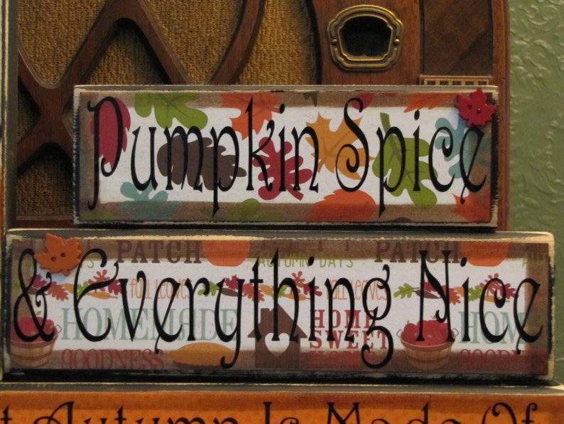 Puede incluir: Dos letreros de madera con dise&ntilde;os de oto&ntilde;o. El letrero superior dice "Pumpkin Spice" en letras negras con un patr&oacute;n de hojas rojas y verdes. El letrero inferior dice "& Everything Nice" en letras negras con un patr&oacute;n de hojas marrones, naranjas y verdes.