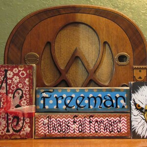 May include: Three wooden blocks with painted designs. The first block has a red and white floral pattern with the letter "M" in black. The second block is blue with white polka dots and the word "Freeman" in black. The third block has a red and white chevron pattern with the words "Always Fall Forward" in black. The last block has a black background with a white and yellow eagle head.